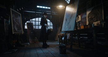 Movie still from “Wednesday” (2022), created by Miles Millar – A man in a black coat and a hat standing in front of an easel; Wide shot, Low angle