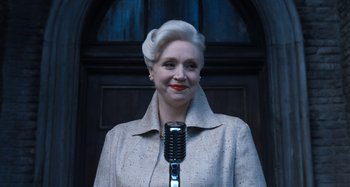 Movie still from “Wednesday” (2022), created by Miles Millar – An older woman is holding a microphone in her hands; Medium shot, Low angle