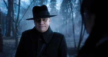 Movie still from “Wednesday” (2022), created by Miles Millar – A man wearing a hat and a coat in the woods; Close Up shot, Over the shoulder angle