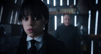 Movie still from “Wednesday” (2022), created by Miles Millar – A woman in a suit and tie standing next to a man in a suit and tie; Close Up shot, Over the shoulder angle
