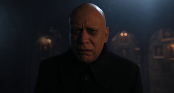 Movie still from “Wednesday” (2022), created by Miles Millar – A bald man in a black shirt is looking at the camera; Close Up shot, Low angle