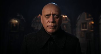 Movie still from “Wednesday” (2022), created by Miles Millar – A bald man in a black coat is looking at the camera; Close Up shot, Low angle