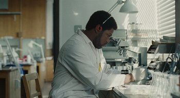 Movie still from “Small Axe” (2020), directed by Steve McQueen – A man working in a lab with a microscope; Medium shot, Low angle