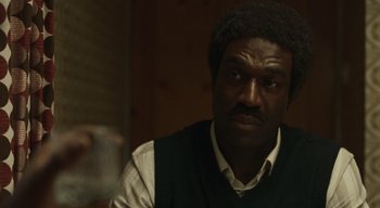 Movie still from “Small Axe” (2020), directed by Steve McQueen – A person sitting at a table holding a glass; Close Up shot, Over the shoulder angle