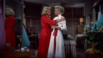 Movie still from “White Christmas” (1954), directed by Michael Curtiz – Two women in red and a white dress one is tying a red ribbon around the neck of the other woman; Medium shot, Low angle