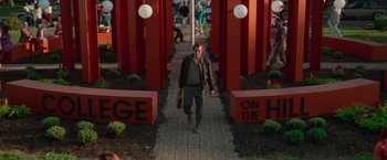 Movie still from “White Noise” (2022), directed by Noah Baumbach – A man walking down a brick path in front of red pillars; Wide shot, High angle