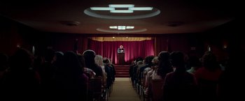Movie still from “White Noise” (2022), directed by Noah Baumbach – A man standing in front of an audience in front of a red curtain; Extreme Wide shot, High angle