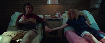 Movie still from “White Noise” (2022), directed by Noah Baumbach – A man and a woman holding hands while sitting on a couch; Medium shot, High angle