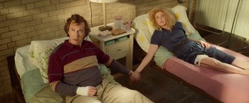 Movie still from “White Noise” (2022), directed by Noah Baumbach – A man and a woman holding hands while sitting on a bed; Medium shot, High angle