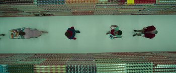 Movie still from “White Noise” (2022), directed by Noah Baumbach – Two people sitting on the ground in a room full of soda cans; Extreme Wide shot, Overhead angle