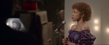 Movie still from “Whitney Houston: I Wanna Dance with Somebody” (2022), directed by Kasi Lemmons – A woman in a purple dress speaking into a microphone; Medium shot, Over the shoulder angle