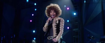 Movie still from “Whitney Houston: I Wanna Dance with Somebody” (2022), directed by Kasi Lemmons – A woman in a black dress and a brown and white jacket; Medium shot, Over the shoulder angle