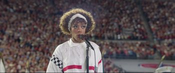 Movie still from “Whitney Houston: I Wanna Dance with Somebody” (2022), directed by Kasi Lemmons – A woman in white shirt holding a microphone on a stage; Medium shot, Low angle