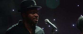 Movie still from “Whitney Houston: I Wanna Dance with Somebody” (2022), directed by Kasi Lemmons – A man in a black hat is speaking into a microphone; Close Up shot, Low angle