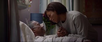 Movie still from “Whitney Houston: I Wanna Dance with Somebody” (2022), directed by Kasi Lemmons – An older man and a young woman laying in a hospital bed; Medium shot, High angle