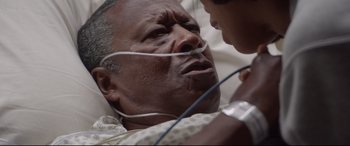 Movie still from “Whitney Houston: I Wanna Dance with Somebody” (2022), directed by Kasi Lemmons – An older man is in a hospital bed with a young boy; Close Up shot, High angle