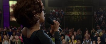 Movie still from “Whitney Houston: I Wanna Dance with Somebody” (2022), directed by Kasi Lemmons – A woman holding a microphone in front of a crowd of onlookers; Medium shot, Over the shoulder angle