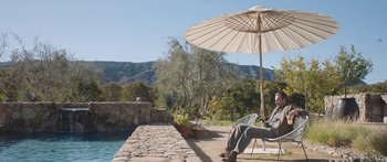 Movie still from “Windfall” (2022), directed by Charlie McDowell – A man sitting under an umbrella next to a swimming pool; Wide shot, High angle