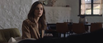 Movie still from “Windfall” (2022), directed by Charlie McDowell – A beautiful young lady sitting at a table in front of a fireplace; Close Up shot, Over the shoulder angle