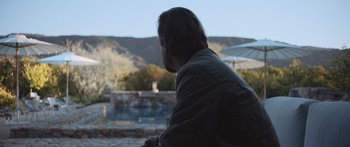 Movie still from “Windfall” (2022), directed by Charlie McDowell – A man sitting in the shade of an umbrella; Medium shot, Over the shoulder angle