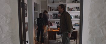 Movie still from “Windfall” (2022), directed by Charlie McDowell – Three people standing in a living room with a bookshelf; Wide shot, Over the shoulder angle