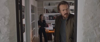 Movie still from “Windfall” (2022), directed by Charlie McDowell – A man and a woman standing in a living room; Medium shot, Over the shoulder angle