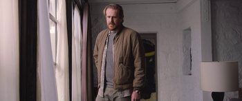 Movie still from “Windfall” (2022), directed by Charlie McDowell – A man standing in front of a window wearing a brown jacket; Medium shot, Low angle