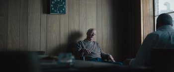 Movie still from “Wormwood” (2017), directed by Errol Morris – An older man sitting in front of a wall clock; Medium shot, Over the shoulder angle