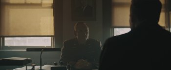 Movie still from “Wormwood” (2017), directed by Errol Morris – A man in a military uniform sitting at a table in front of a portrait; Medium shot, Over the shoulder angle