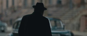 Movie still from “Wormwood” (2017), directed by Errol Morris – A man wearing a hat and a black coat walking down the street; Close Up shot, Over the shoulder angle