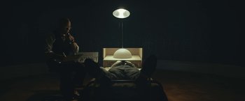 Movie still from “Wormwood” (2017), directed by Errol Morris – A man laying down under a lamp in a dark room; Wide shot, High angle