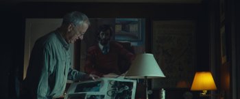 Movie still from “Wormwood” (2017), directed by Errol Morris – An older man looking at a book in a dark room; Medium shot, Over the shoulder angle