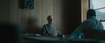 Movie still from “Wormwood” (2017), directed by Errol Morris – An older man sitting in a chair in front of a clock; Medium shot, Over the shoulder angle