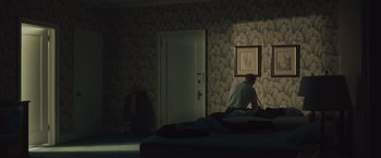 Movie still from “Wormwood” (2017), directed by Errol Morris – A man sitting on a bed in front of a painting; Wide shot, Low angle