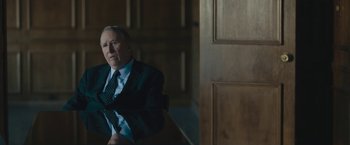 Movie still from “Wormwood” (2017), directed by Errol Morris – A man in a suit and tie sitting in front of a wooden door; Medium shot, High angle