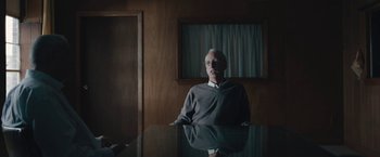 Movie still from “Wormwood” (2017), directed by Errol Morris – A man standing in front of a table in a room; Medium shot, Low angle