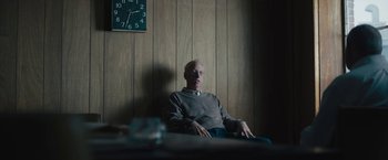 Movie still from “Wormwood” (2017), directed by Errol Morris – An older man sitting in a chair in front of a clock; Medium shot, Over the shoulder angle