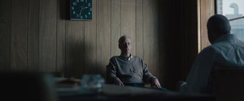 Movie still from “Wormwood” (2017), directed by Errol Morris – An older man sitting in front of a wall clock; Medium shot, Low angle