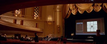 Movie still from “Steve Jobs” (2015), directed by Danny Boyle – People are sitting on the stage of a large theater; Extreme Wide shot, High angle