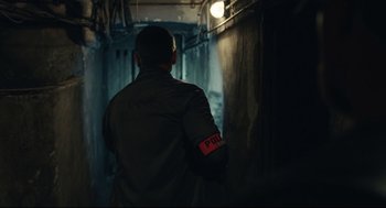 Movie still from “Stillwater” (2021), directed by Tom McCarthy – A man with a police arm band is standing in a dark room; Medium shot, Over the shoulder angle