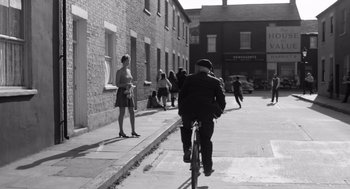 Movie still from “Belfast” (2021), directed by Kenneth Branagh – A man riding a bike down a street past a brick building; Wide shot, Over the shoulder angle