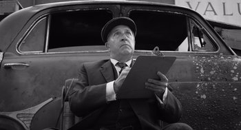 Movie still from “Belfast” (2021), directed by Kenneth Branagh – An older man holding a clipboard while sitting next to an open trunk; Medium shot, Low angle