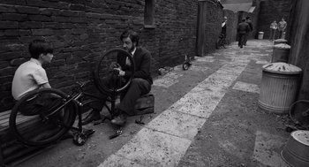 Movie still from “Belfast” (2021), directed by Kenneth Branagh – A man sitting next to a bicycle on the side of the road; Wide shot, High angle