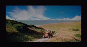 Movie still from “Belfast” (2021), directed by Kenneth Branagh – An old car is driving down a dirt road through a grassy field; Extreme Wide shot, High angle