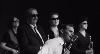 Movie still from “Belfast” (2021), directed by Kenneth Branagh – A man holding a microphone in front of two other people; Medium shot, Over the shoulder angle