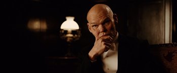 Movie still from “The Assassination of Jesse James by the Coward Robert Ford” (2007), directed by Andrew Dominik – A bald man in a tuxedo is looking at the camera with his hand under his chin; Close Up shot, Low angle