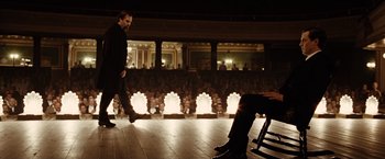 Movie still from “The Assassination of Jesse James by the Coward Robert Ford” (2007), directed by Andrew Dominik – A man standing on a stage in front of a chair; Wide shot, Over the shoulder angle