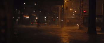 Movie still from “The Batman” (2022), directed by Matt Reeves – A city street at night with a person on a scooter on the sidewalk; Extreme Wide shot, High angle