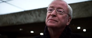 Movie still from “The Dark Knight” (2008), directed by Christopher Nolan – An older man wearing glasses and a black jacket; Close Up shot, Low angle