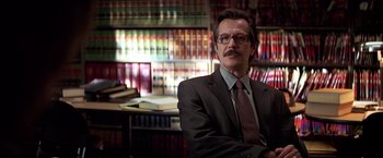 Movie still from “The Dark Knight” (2008), directed by Christopher Nolan – A man in a suit sitting in front of a book shelf; Close Up shot, Low angle